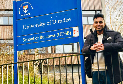: A student poses in front of a sign for the University of Dundee, smiling, wearing a black jacket and light coloured jumper