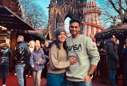 A young couple posing together at a busy Christmas market, male wearing a grey jumper and female wearing a cream jumper with a hat.