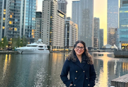Jeanett stands next to a canal with a yacht in the background and tall high rise buildings on each side of the water