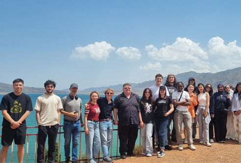 A large group of students, standing in a line smiling at the camera, standing outside with a lake and mountains behind them