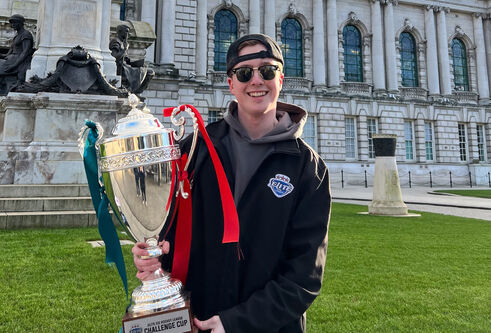 Student wearing a black jacket, grey hoodie, backwards cap, sun glasses and smiling at the camera holding a trophy outside