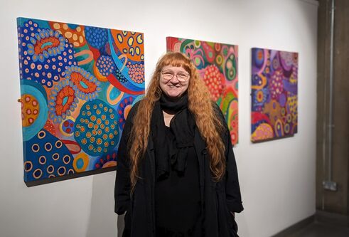 Artist Janice Aikten stands smiling in front of 3 of her colourful canvases