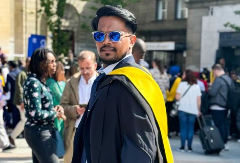 Mohammed Suhail wears sunglasses and graduation robes with a graduation yellow sash in a crowd of people