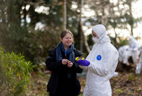 One person in white ppe and a face mask holds a yellow device showing it to someone, there are trees around them.