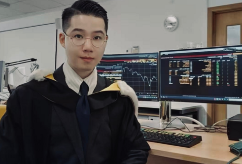 Student sitting infront of a computer, with a graph and statics on the screen, wearing his graduation gown, white shirt and glasses.