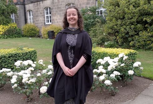 woman standing on garden path with flowers and grass behind her and an older looking house in the background. She is wearing a graduation gown and smiling at the camera