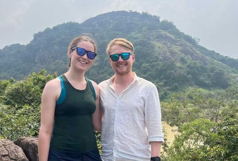 Two people wearing sunglasses stand in front of a mountain