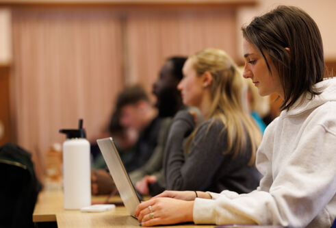 Side view of a young woman sat at a desk with a laptop open looking a the screen. Others sat next to her also look to be working