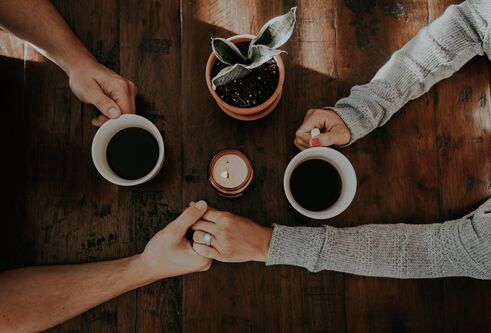 A top-down image of two people hold hands over a table. Their other hands are holding coffee cups.