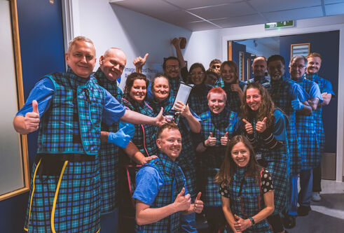 A group of people wearing blue and tartan scrubs stand in a corridor with white walls and blue doors.