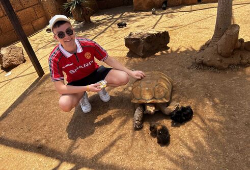 Aidan Harper ewars a red football top, shorts and a cap with sunglasses. He squats down to pet a tortoise, there are also guinea pigs.