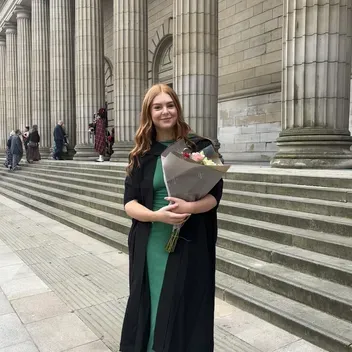 A person stands on the steps outside of a building with large pillars, they are wearing graduation robes and a green dress, holding a bunch of flowers.