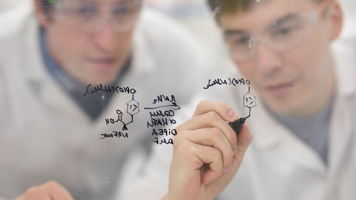 A lecturer and student in front of a perplex glass writing an equation in black pen