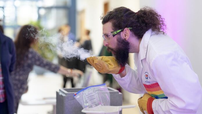 A man wearing goggles and a rainbow coloured lab coat blows a smoky gas from the gloved hand in a white-walled gallery space