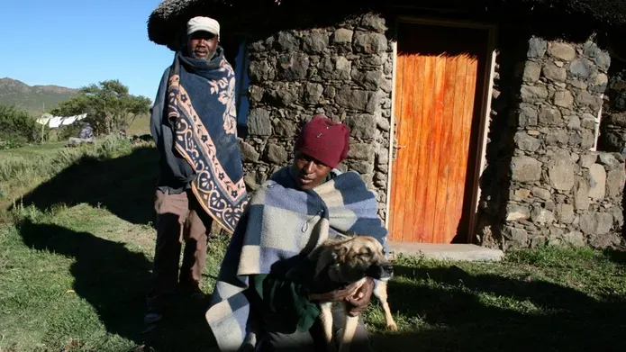 2 people and a dog sitting outside of a stone house with a wooden door