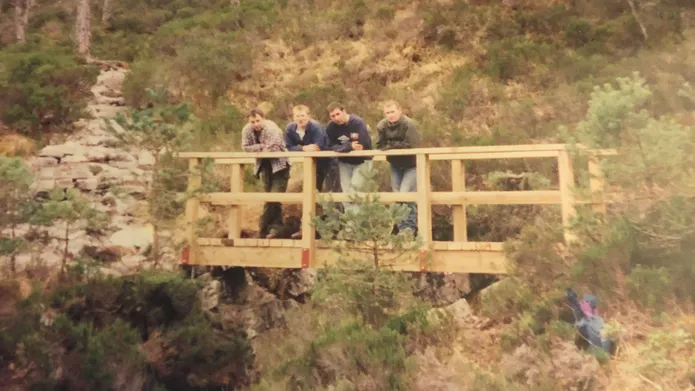 Four men in their twenties lean against the handrail on a freshly built, unpainted, wooden bridge. The bridge crosses a small rocky gorge surrounded by foliage and further rock formations.