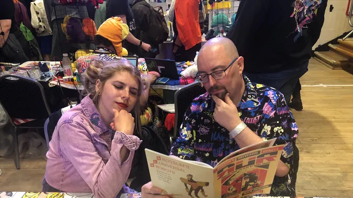 Two people sit at a table covered with comics, both looking together at an open comic with their hands on their chins.