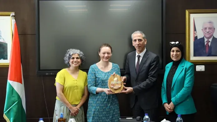 Jane Macaskill stands holding an award, surrounded by three Palestinian colleagues. They are standing in an office space with the flag of Palestine hanging beside a desk.