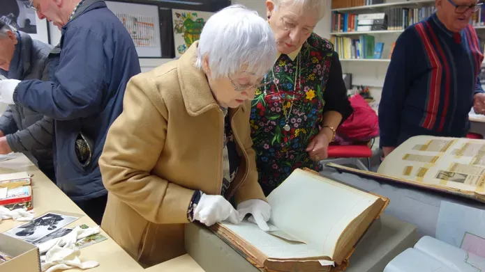 People are standing around a set of tables, a woman in the middle is leaning over looking at a book.