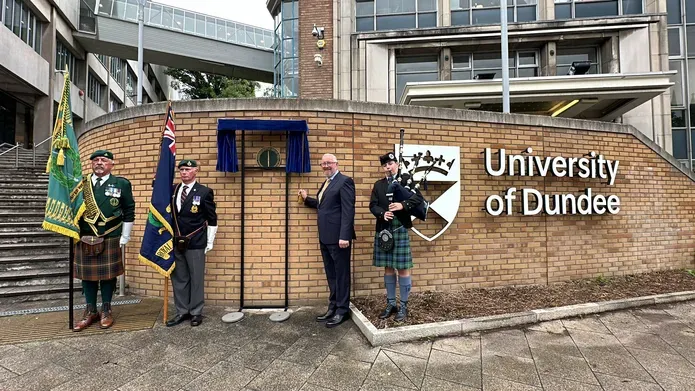 Two men in formal wear holding flags, one man in a suit standing next to a plaque and a piper