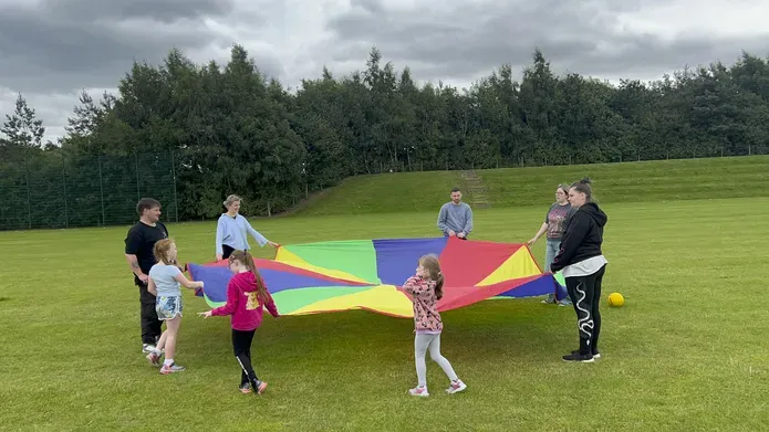 A group of adults and children stand in a circle holding the edges of a rainbow coloured parachute, in a field with trees in the background.