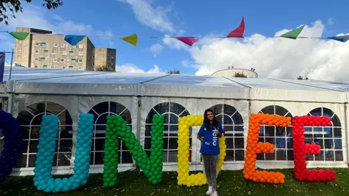 A person stands in front of a white marquee with colourful bunting flying overhead. There is large writing spelling out "DUNDEE" with balloons.