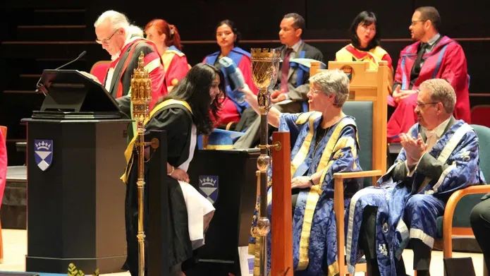 Anju Matthew on stage at a graduation ceremony, getting bopped on the head with a scroll. People in ceremonial robes are sitting.