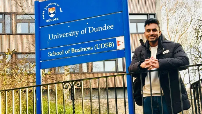: A student poses in front of a sign for the University of Dundee, smiling, wearing a black jacket and light coloured jumper