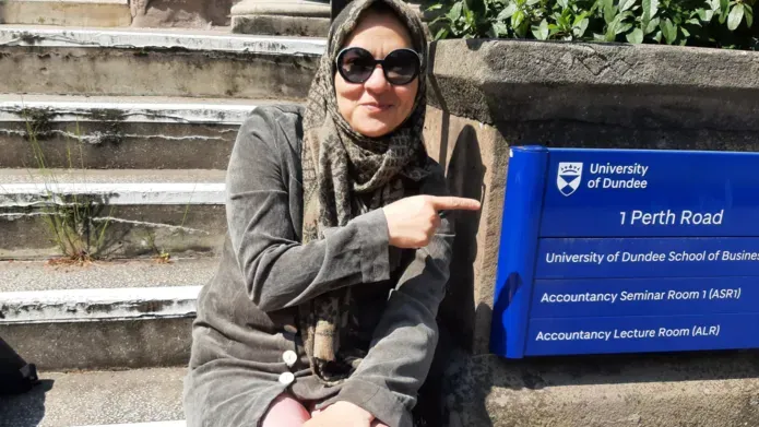 PhD student wearing sunglasses and a headscarf, sitting on the stepsand point at a University sign and address