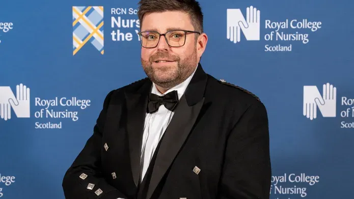 Grant Cooper, in kilt, standing infront of a blue, branded backdrop.