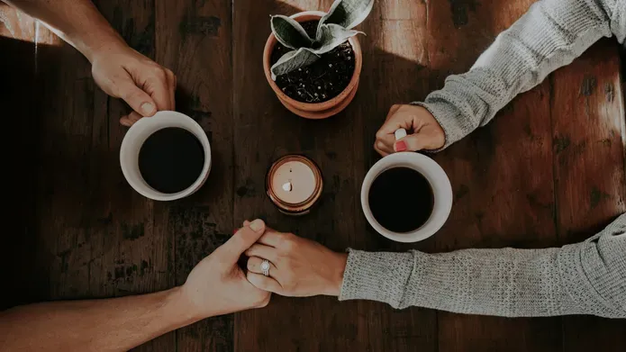 A top-down image of two people hold hands over a table. Their other hands are holding coffee cups.