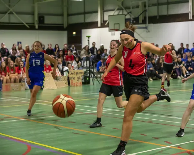 a group of young women in their 20's, some wearing red sporting gear and others blue, running on a basketball pitch chasing a ball