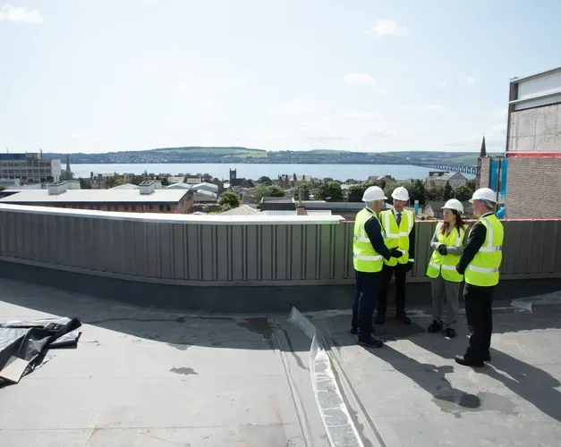 A group of people in high vis vests are stood on a roof in a group speaking to eachother. There is a beautiful backdrop showing the river tay behind them