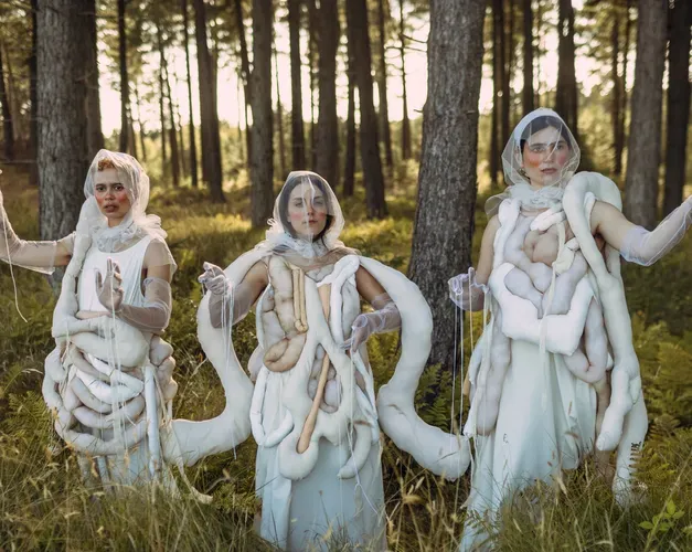 Three women wearing 3D fabric body parts representing the human gut on their outfits with their arms up dancing in a forest