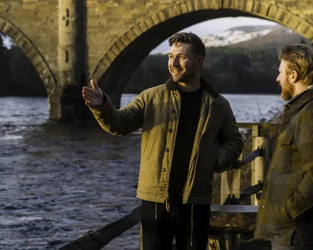 Man standing next to river, gesturing, with bridge in the background