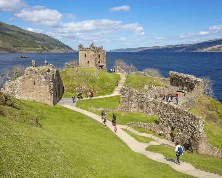 Urquhart Castle near Inverness. People walk around the ruins on a bright summer's day. Behind the castle are some hills and an expanse of water