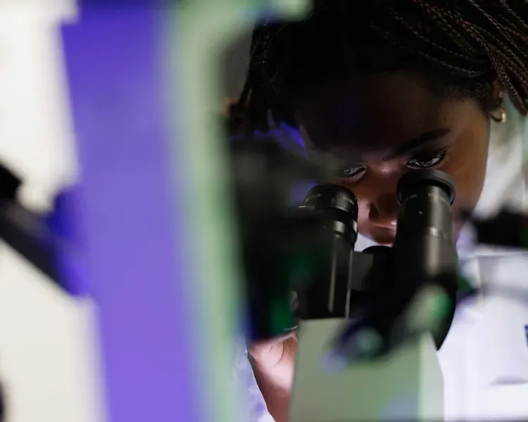 A woman in a lab coat looks through a microscope. A blue-green line cuts across the image.