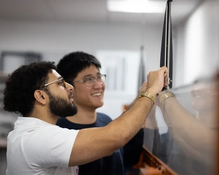Two people stand together in front of a large piece of glass, one of them holds a device against it.
