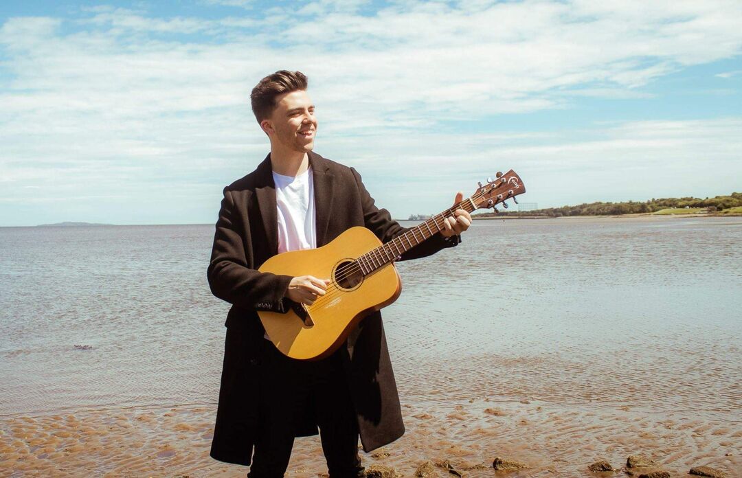 Lewis Kennedy holding a guitar on a beach