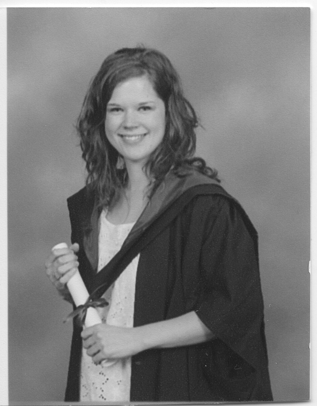 Sheryl Bartlett stands in graduation robes holding her graduation certificate scroll.