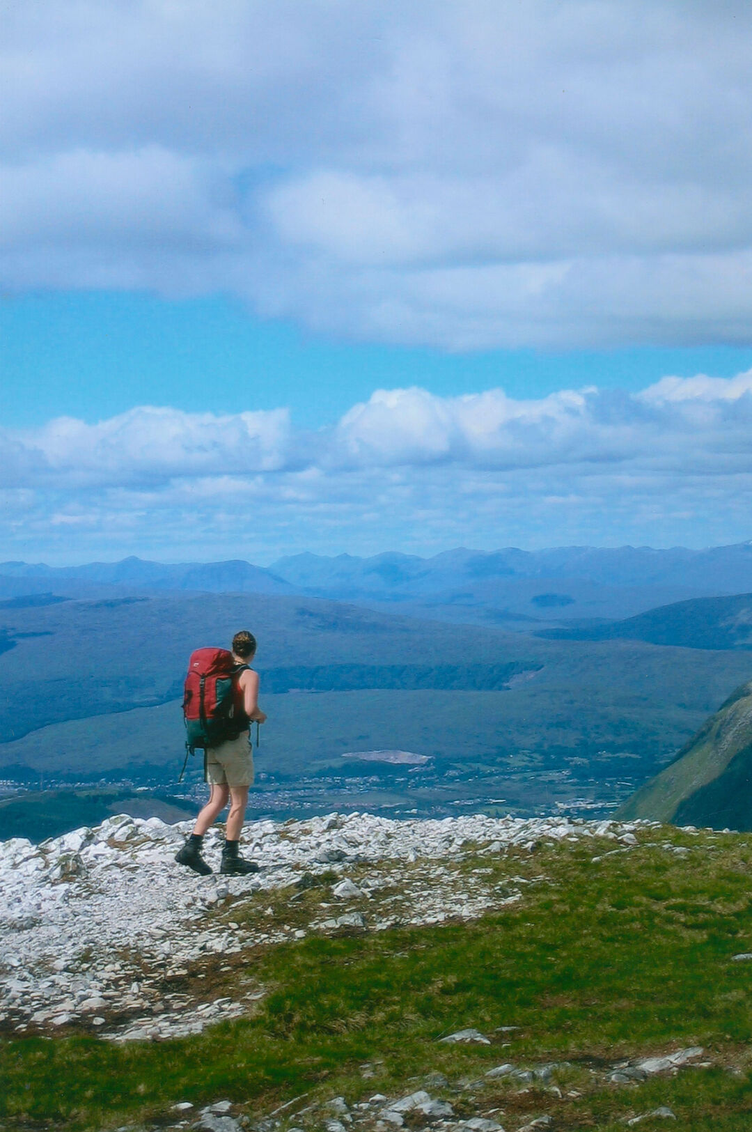 Sheryl Bartlett carries a red backpack and stands on top of a mountain facing out across the vista.