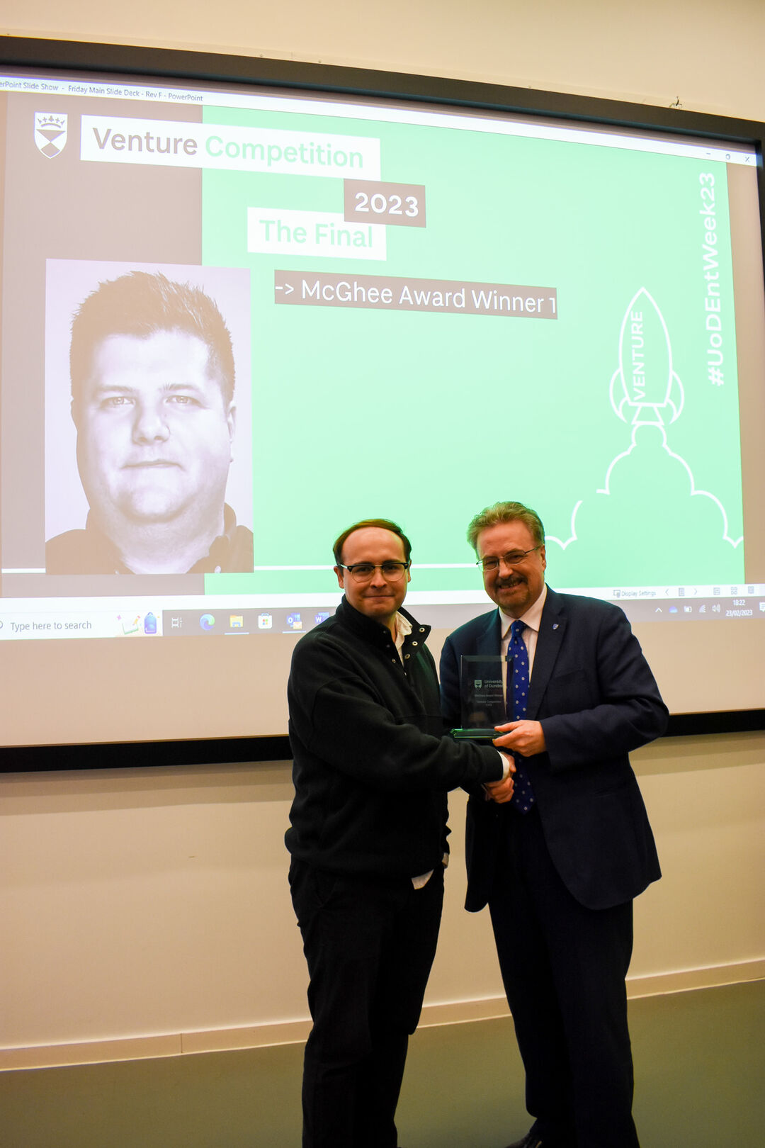 A man standing in front of a projector screen, shaking hands with another man.