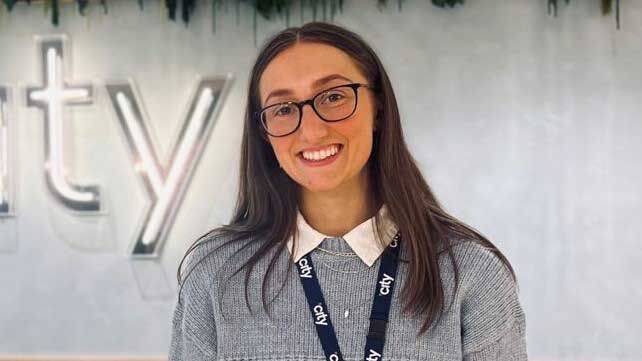 A girl with long brown hair and glasses, wearing a grey sweatshirt with a blue lanyard round her neck.