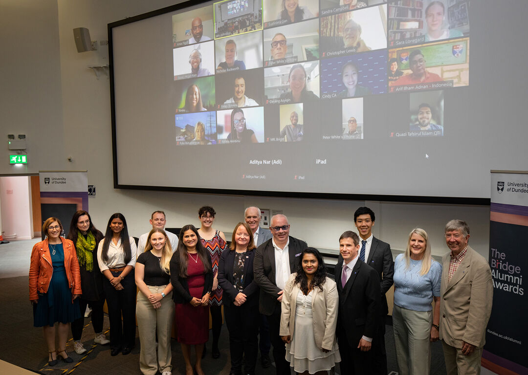 A group photo of Alumni who attended the award ceremony with a huge screen in the background of alumni on Zoom