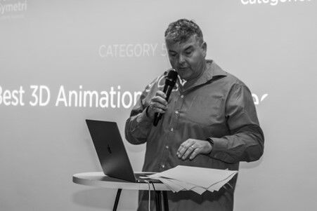 A man stands giving a presentation. He is holding a microphone and looking down at a laptop and sheets of paper.