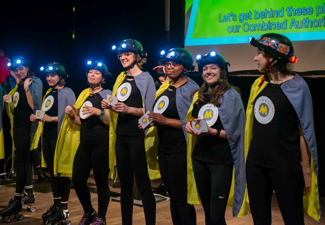 The cast of Bus Regulation production on stage wearing capes and hard hats