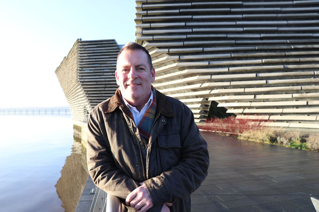 A man in a brown jacket stood outside with his hands together, in ftont of the V&A museum, leaning on a railing 