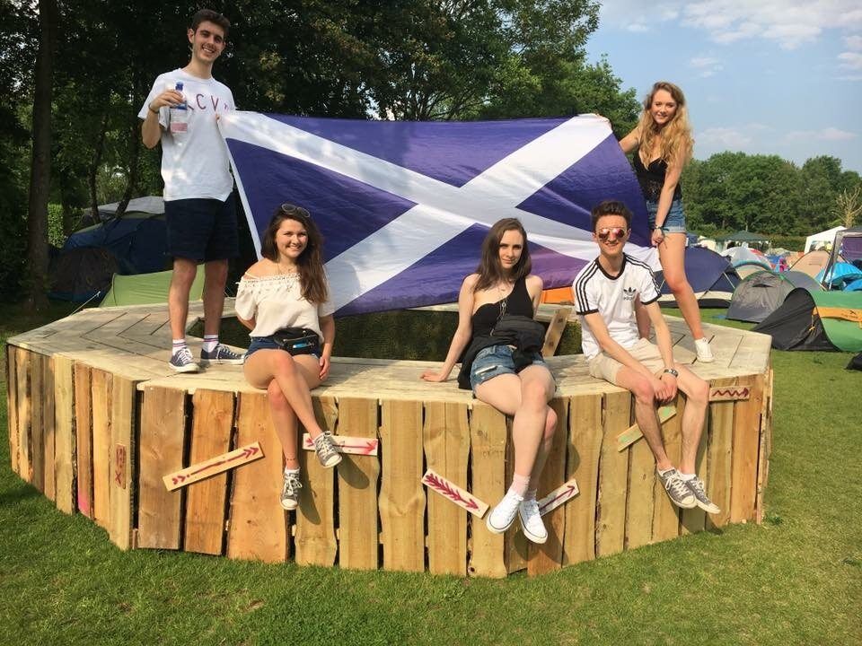 Five people sat and stood on a wooden structure. Two people at the back hold the Scottish flag up while three other people sit in front of it