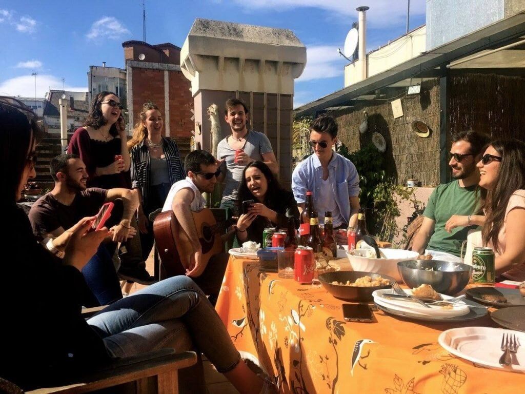 A group dinner. A sunny scene with a large number of people around a table with drinks and plates. One person has an acoustic guitar