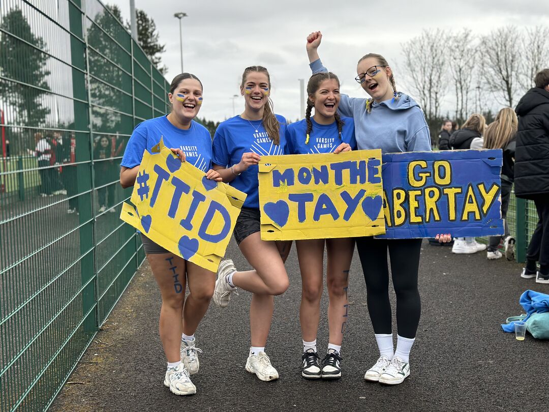 group of girls wearing blue and yellow sports outfits, holding signs of support for Abertay